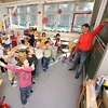 Lehrer vor einer Grundschulklasse, alle Kinder bewegen die Arme.