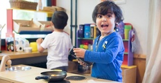 Ein dunkelhaariges Kind mit blauem Pullover und einer Dose in der Hand in einer Spielküche. Vor ihm steht eine kleine Bratpfanne. Im Hintergrund ein Kind von hinten mit weißem T Shirt.