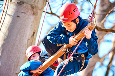 2 Jungs mit rotem Helm und blauer Kleidung beim Klettern mit Seilen und Holzstange an einem großen Baum