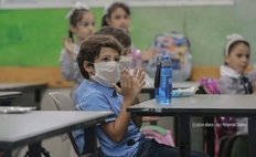 Klatschendes Kind mit Maske und hellblauem Hemd in der Klasse sitzend, unscharf im Hinterrung Mitschülerinnen, vor dem Kind auf dem Tisch blaue Trinkflasche