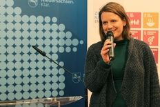 Lächelnde Ministerin in dunkler Strickjacke und Mikro in der Hand in der rechten Bildhälfte stehend, im Hintergrund links blaue Rückwand mit weißen großen Punkten und angeschnitteneem Niedersachsen-Logo oben.