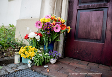 Untere linke Ecke einer dunkelbraunen Holz-Außentür, davor auf dem dunkelroten Steinboden verschiedene bunte Blumensträuße in Vasen und ein Grablicht.