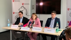 3 Personen an einem hellen Holztisch sitzend ,it Namensschilder davor, im Hintergrund ein schwarzer Monitor, Personen v. l. n. r: Herr Reimann-Lübker, Ministerin Hamburg, Herr Dierkes-Knauer, alle MK