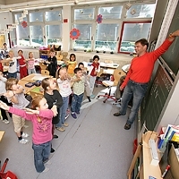 Lehrer vor einer Grundschulklasse, alle Kinder bewegen die Arme.