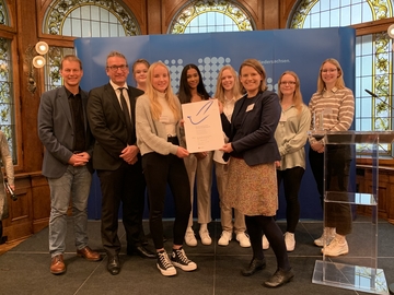 Gruppenfoto auf einer kleinen Bühne mit Ministerin und der Urkunde des Schülerfriedenspreises