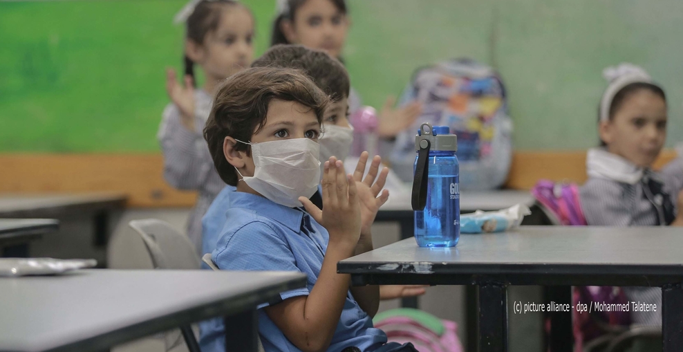 Klatschendes Kind mit Maske und hellblauem Hemd in der Klasse sitzend, unscharf im Hinterrung Mitschülerinnen, vor dem Kind auf dem Tisch blaue Trinkflasche
