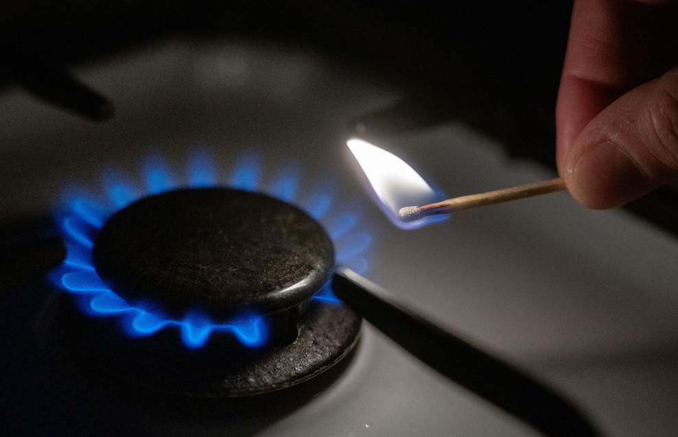 Blau brennende Herd-Gasflamme, daneben ein brennendes Streichholz von zwei Fingerspitzen gehalten