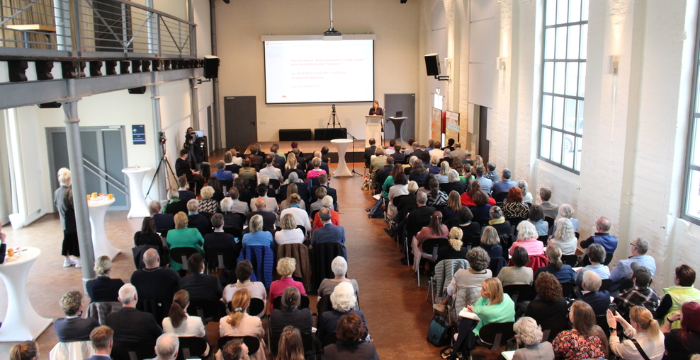 Menschen im Saal, sitzend, eine Frau am Pult trägt vor