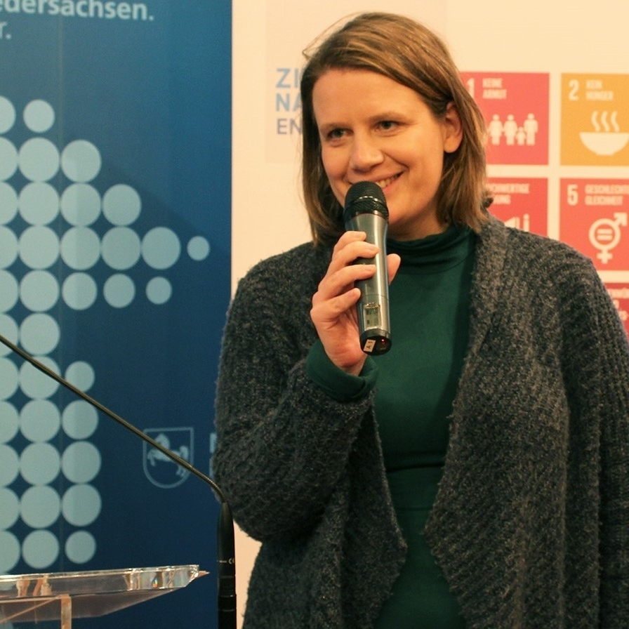 Ministerin in dunkler Strickjacke und Mikro in der Hand in der rechten Bildhälfte stehend, im Hintergrund links blaue Rückwand mit weißen großen Punkten und angeschnitteneem Niedersachsen-Logo oben.