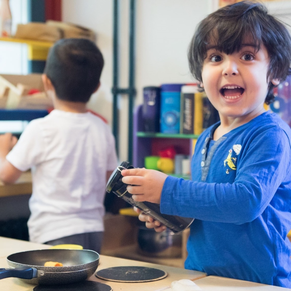 Ein dunkelhaariges Kind mit blauem Pullover und einer Dose in der Hand in einer Spielküche. Vor ihm steht eine kleine Bratpfanne. Im Hintergrund ein Kind von hinten mit weißem T Shirt.