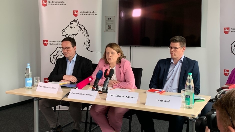 3 Personen an einem hellen Holztisch sitzend ,it Namensschilder davor, im Hintergrund ein schwarzer Monitor, Personen v. l. n. r: Herr Reimann-Lübker, Ministerin Hamburg, Herr Dierkes-Knauer, alle MK